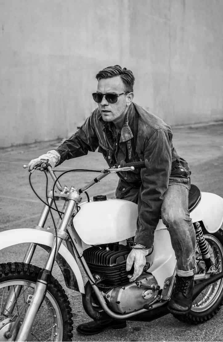 Black and white photograph of Ewan McGregor in sunglasses and a denim jacket riding a vintage motocross bike.