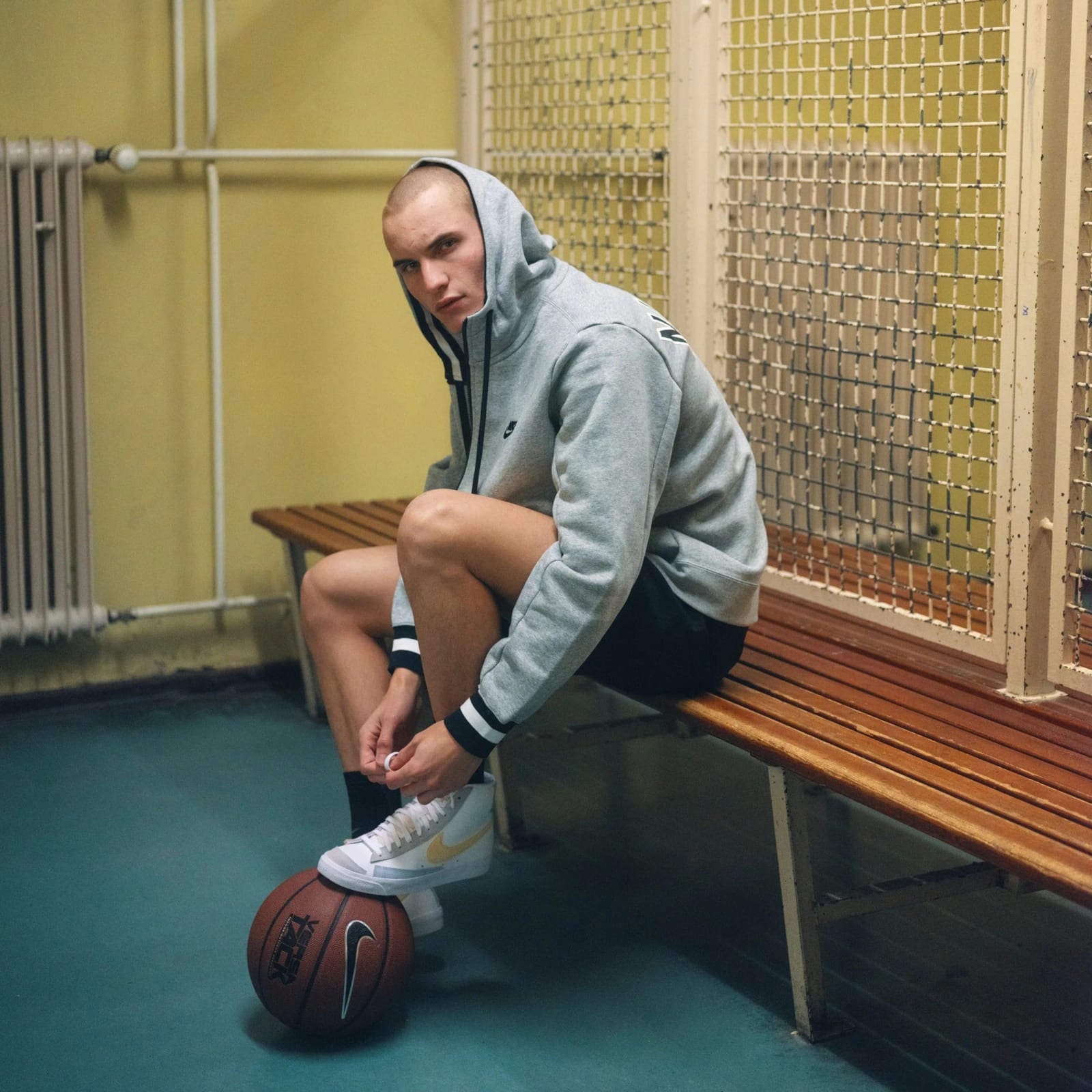 A man in a grey Nike hoodie tying his trainers on a wooden bench in a tiled sports hall, a basketball at his feet.