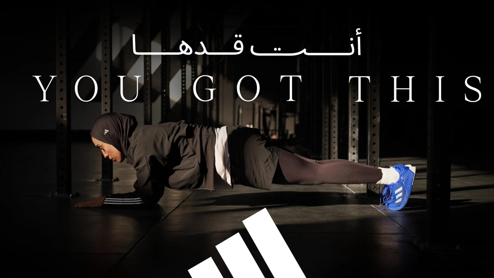 A woman in a black hijab and shorts holding a plank on a gym floor, under the slogan You got this in English and Arabic and the adidas logo.