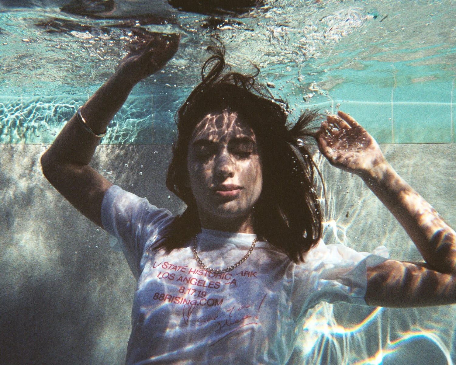 A young woman in a printed white T-shirt floating underwater in a pool, eyes closed, light rippling across her face.