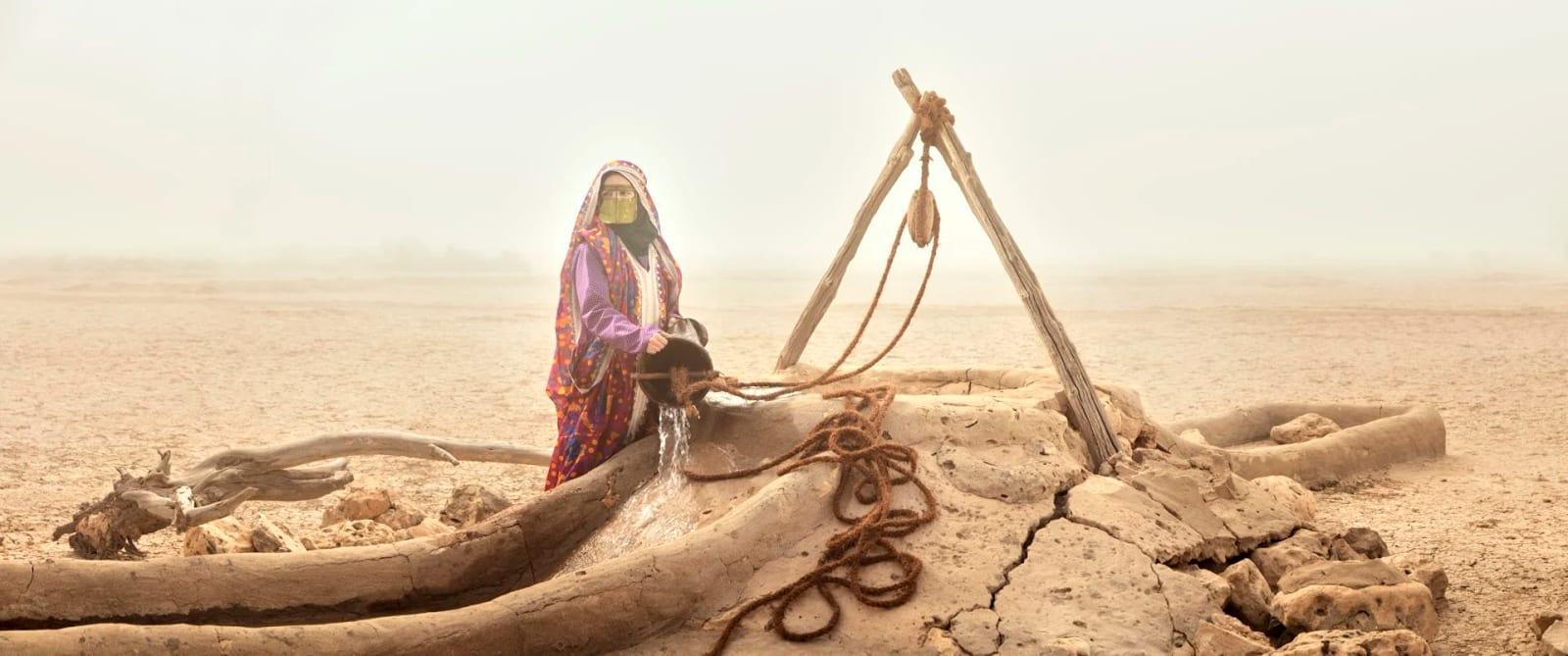 A woman in patterned robes standing at a wooden well frame in a wide empty desert.