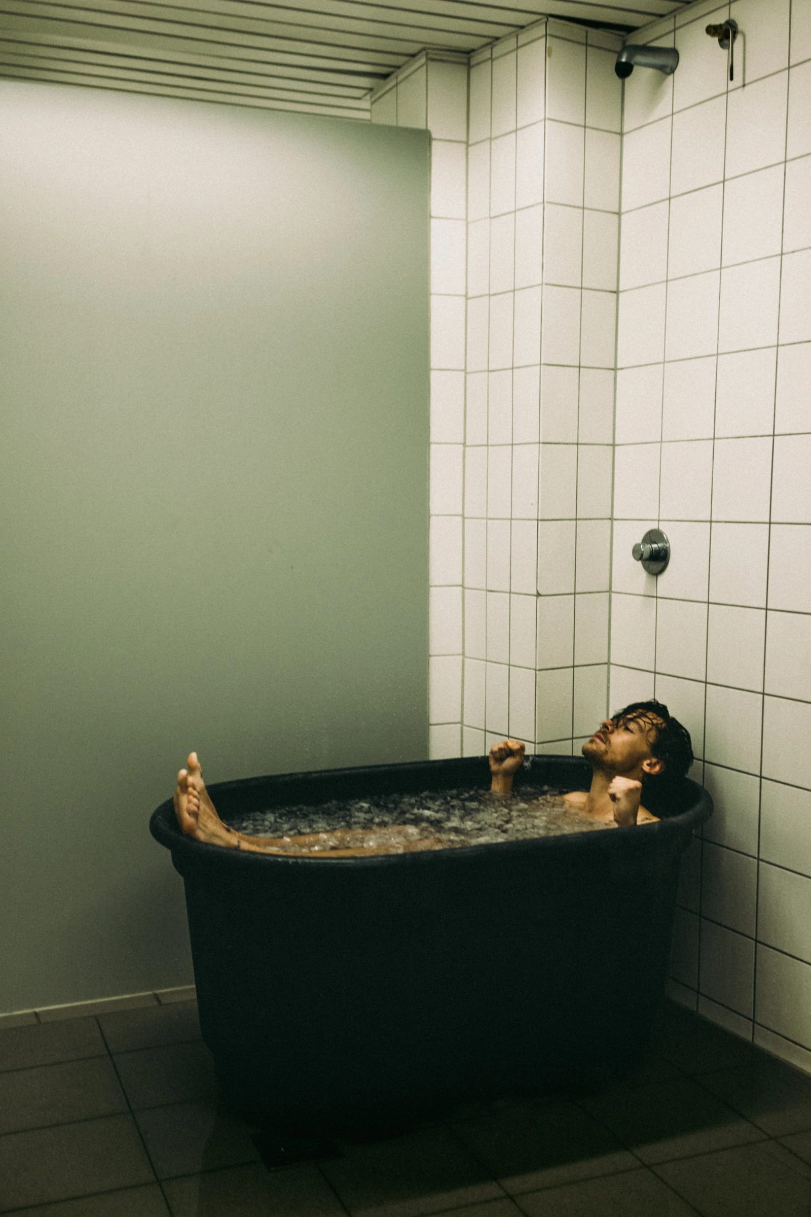 Harry Styles sitting in a black tub of ice water in a white-tiled shower room.