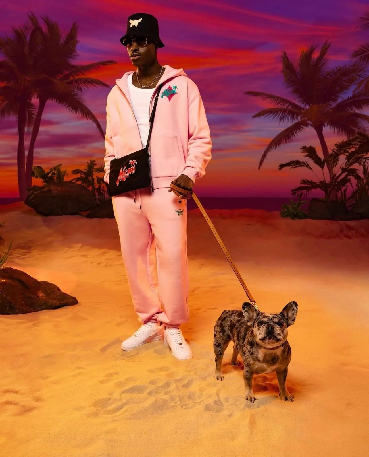 A man in a pink tracksuit and bucket hat walking a French bulldog on a beach set under a purple sunset.