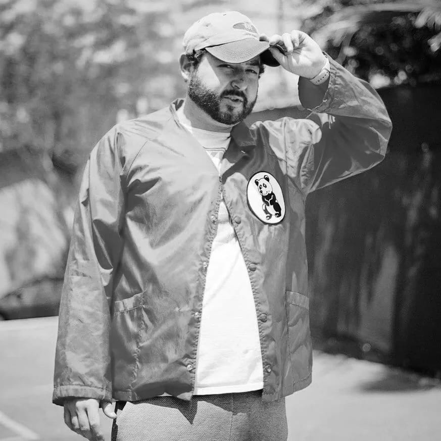 Black and white photograph of Joe Weil in a cap and a jacket with a panda patch, one hand on the brim.