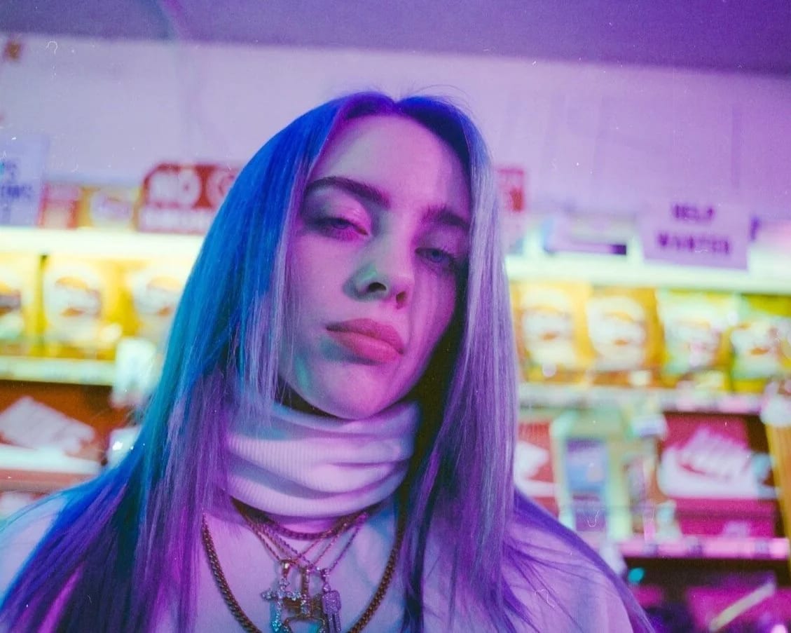 A young woman with long hair lit blue and purple, in a white turtleneck and layered charm necklaces, looking into the camera in a convenience-store aisle.