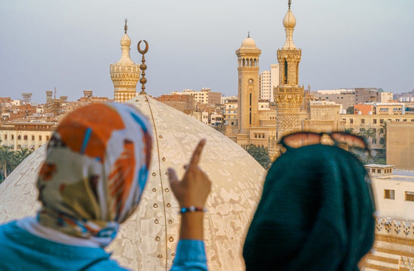 Two women in headscarves looking out over domes and minarets, one pointing across the city.