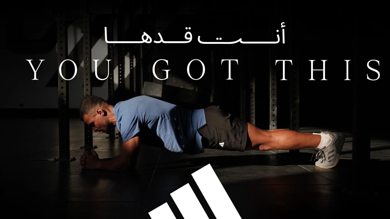 A man in a blue T-shirt holding a plank on a gym floor, under the slogan You got this in English and Arabic and the adidas logo.