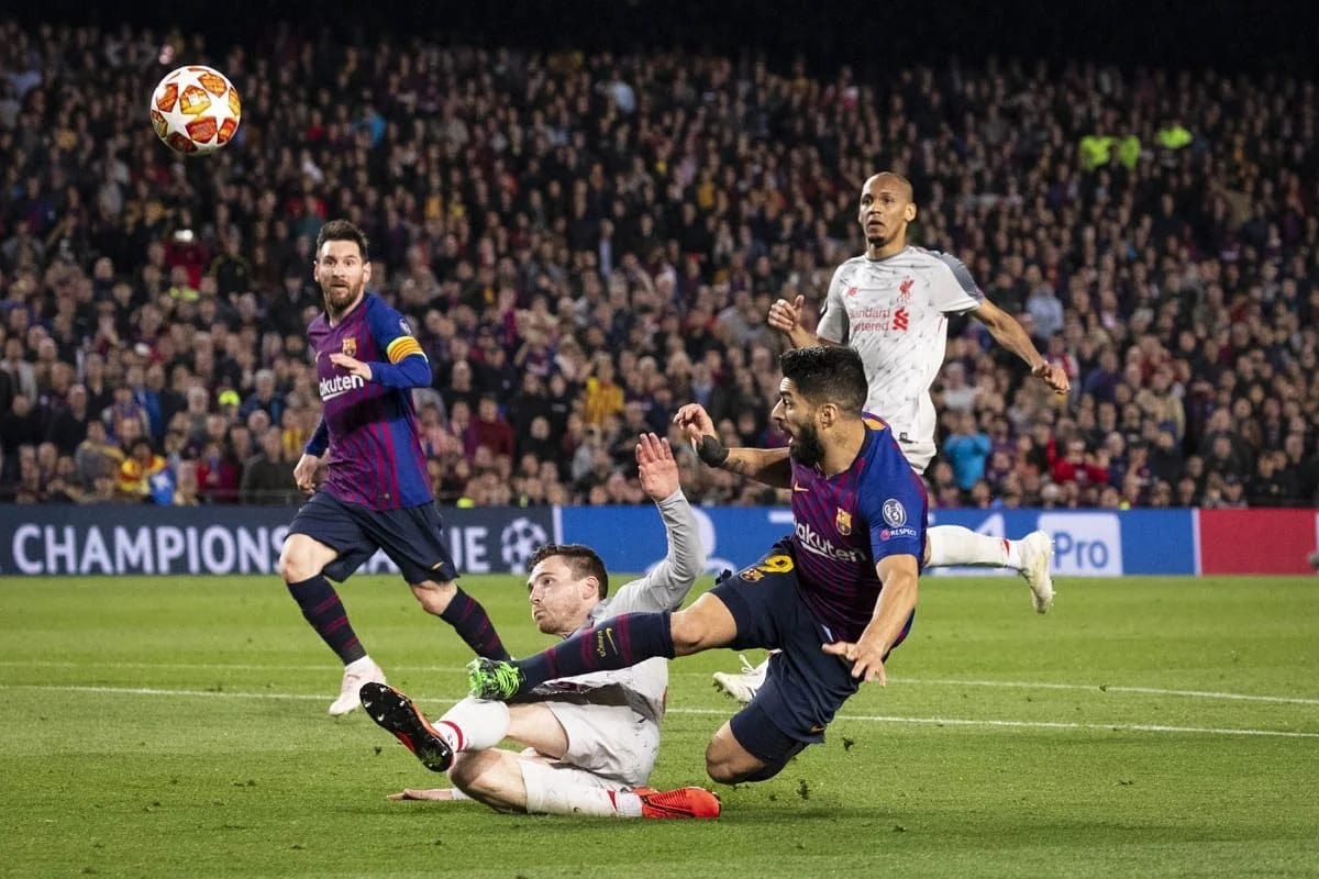 A footballer in blue and red strikes the ball past a sliding defender in white as the crowd watches, a Champions League hoarding behind.