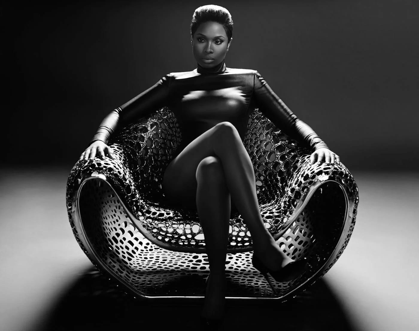 Black and white photograph of a woman in a skin-tight black outfit sitting cross-legged in a sculptural perforated chair.