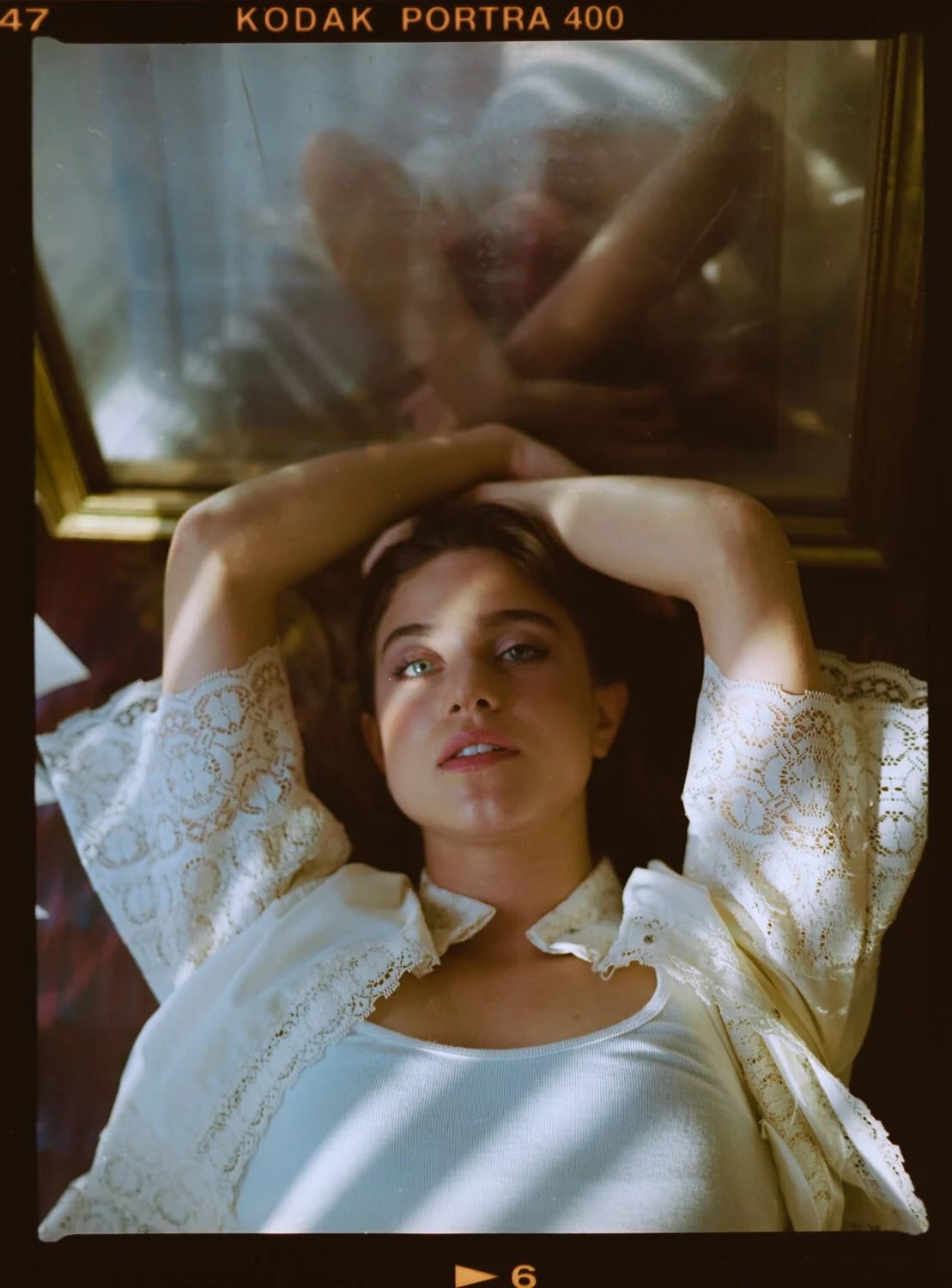 A film frame of a woman in a white lace blouse lying with her arms above her head, her reflection in a mirror behind.