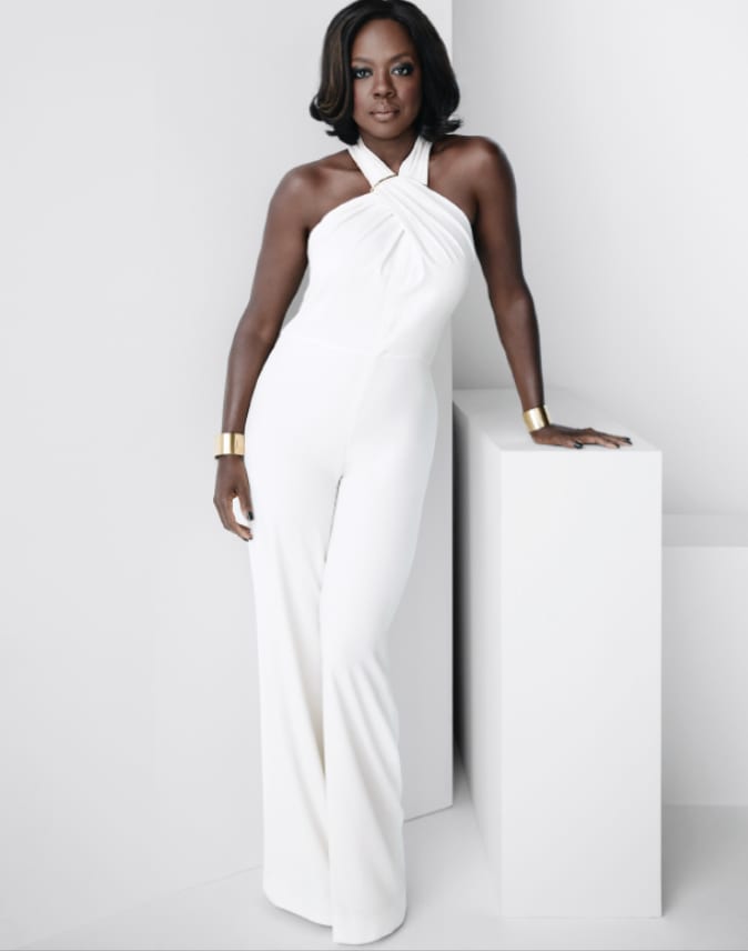 A woman in a white halter-neck jumpsuit and wide gold cuffs, one hand resting on a white plinth against a white background.