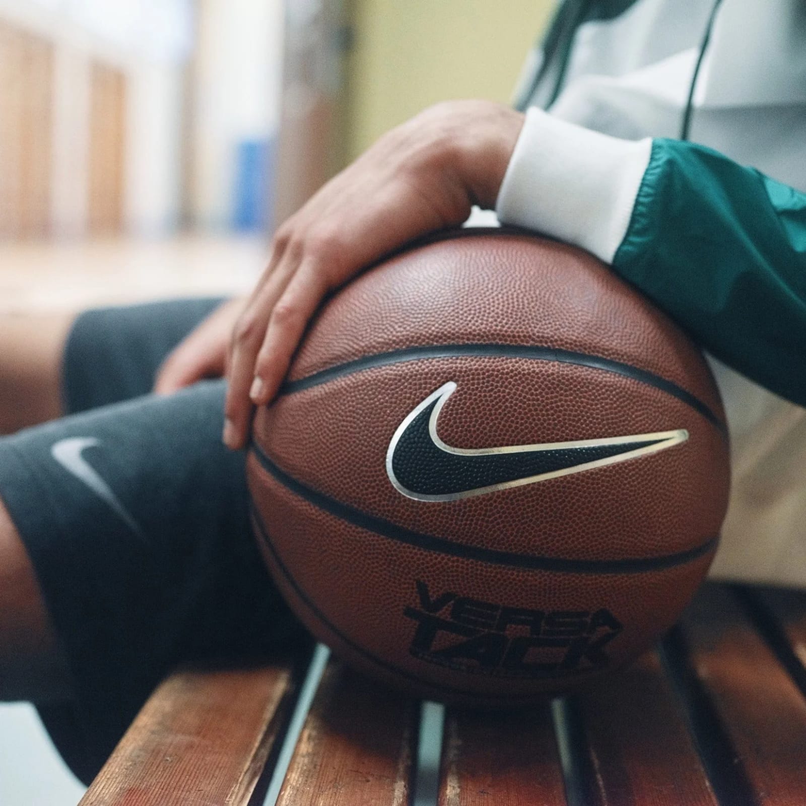 Close-up of a hand resting on a Nike basketball on a wooden bench, the sleeve of a green and white track jacket at the edge of the frame.