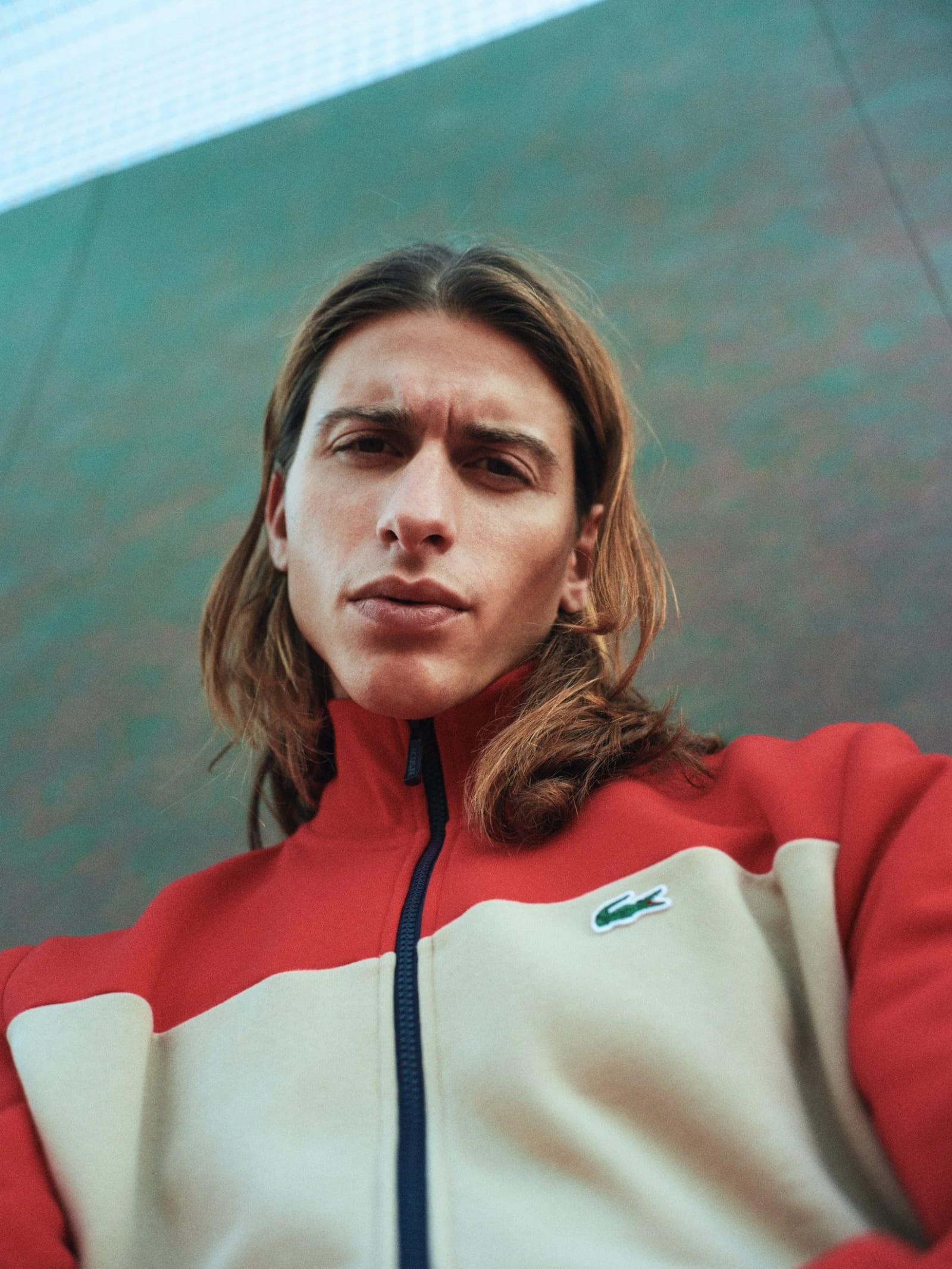 Portrait of a long-haired man in a cream and red Lacoste track jacket against a green court wall.