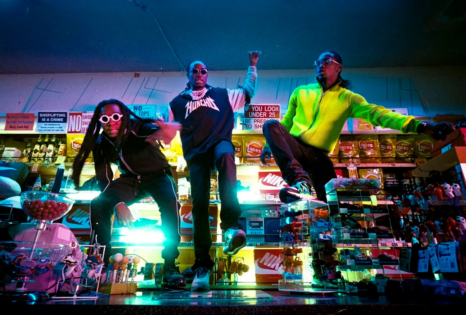 Three men in a brightly lit convenience store at night, one crouching in sunglasses, one standing, one in a neon green jacket climbing the counter.