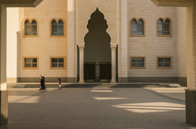 A woman and a child crossing the sunlit courtyard of a sandstone building with an arched doorway.