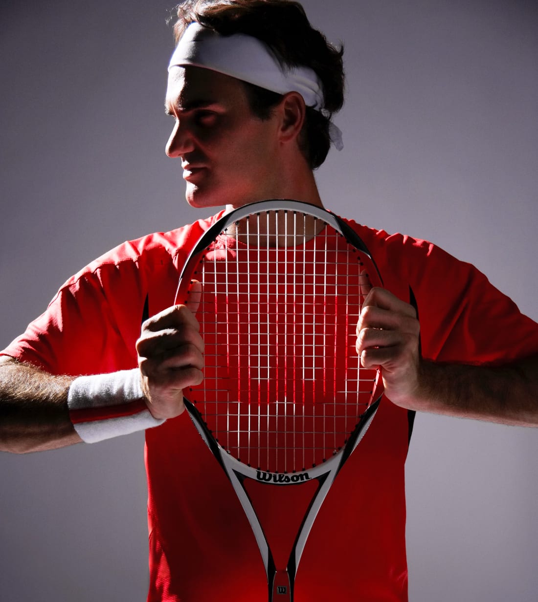 A tennis player in a red shirt and white headband holding a Wilson racket in front of his face.