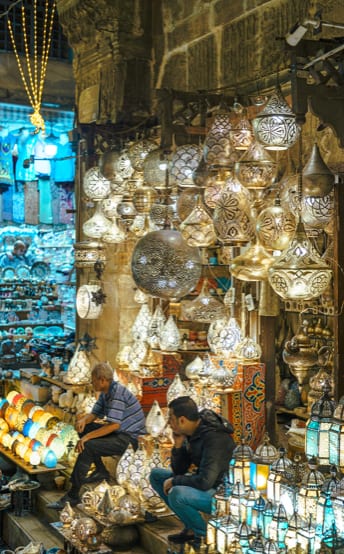 Two men sitting among hundreds of ornate hanging lamps in a market stall at night.