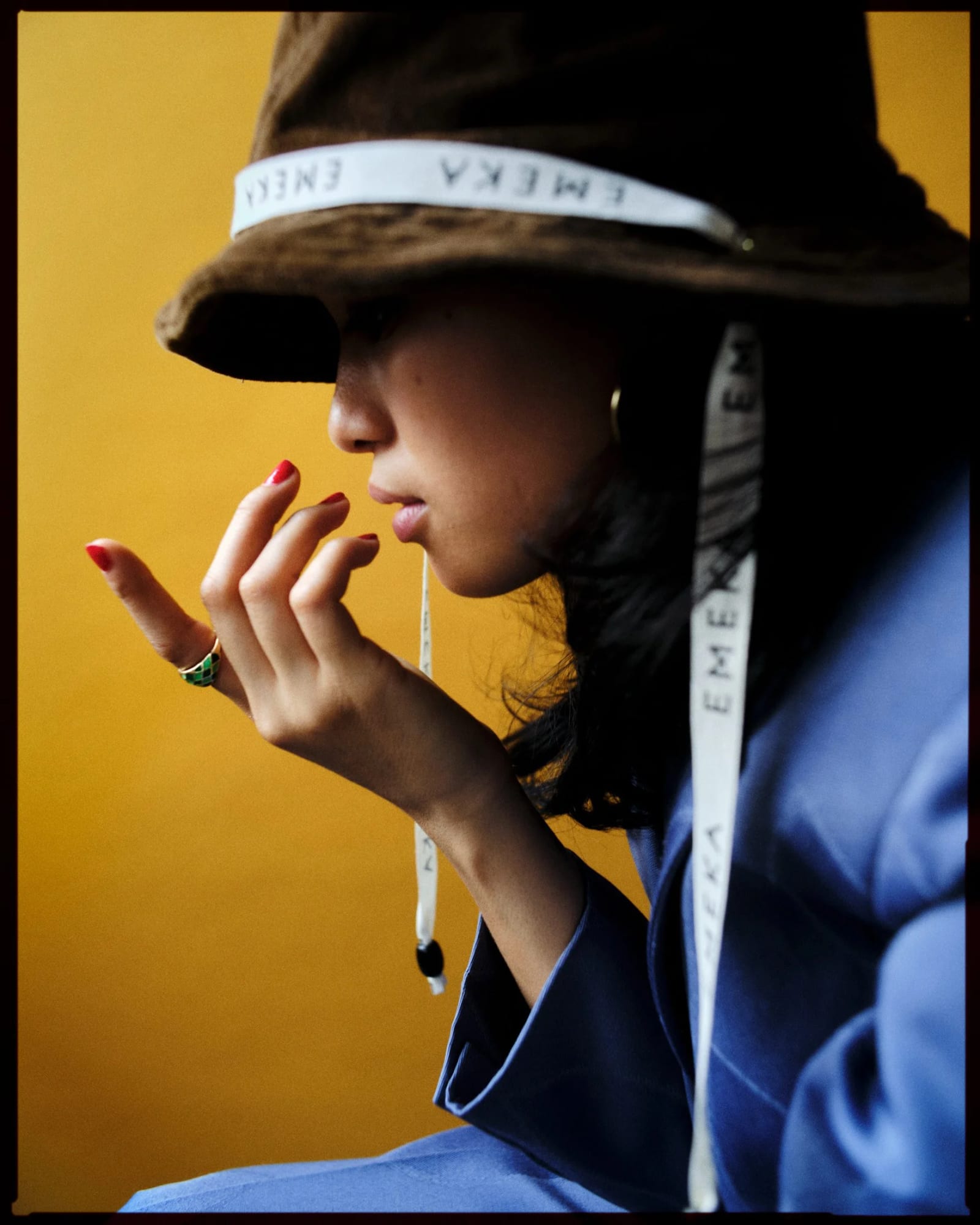 Profile of a woman in a brown bucket hat with a white strap lettered EMEKA, red-nailed fingers raised to her lips, against a yellow wall.