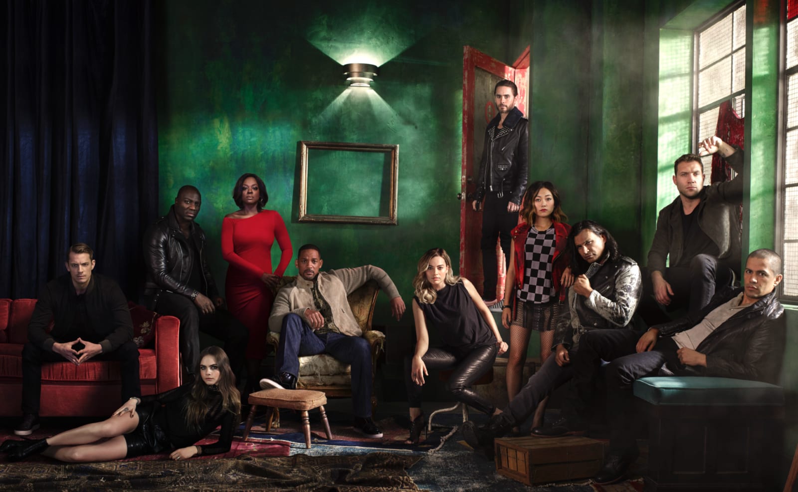 A large cast posed in a dim green-walled room, some seated on red sofas and one standing in a red doorway.