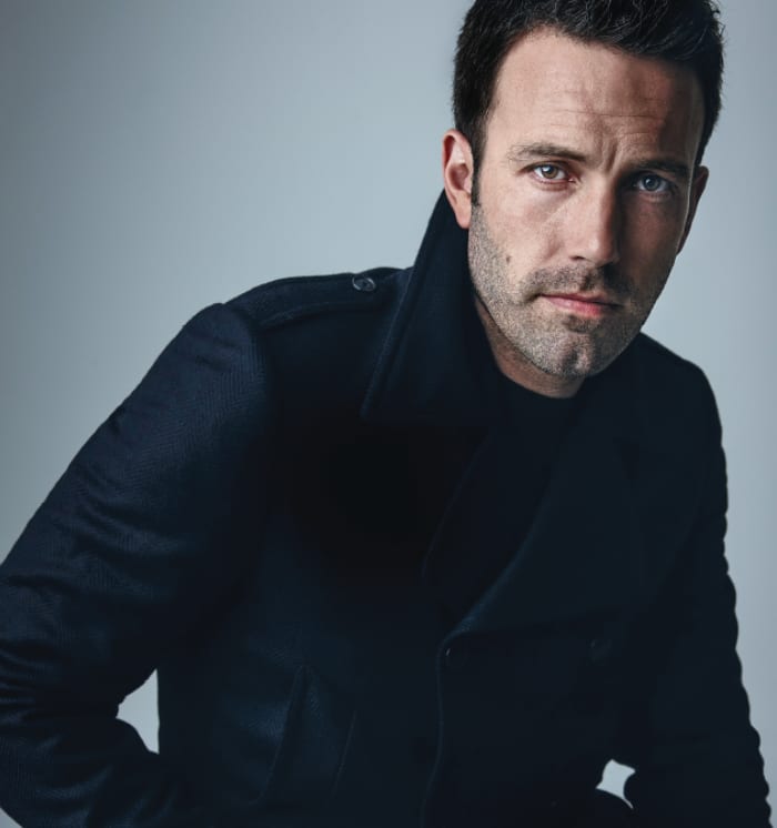 A man with stubble in a dark coat looking at the camera against a cool grey background.