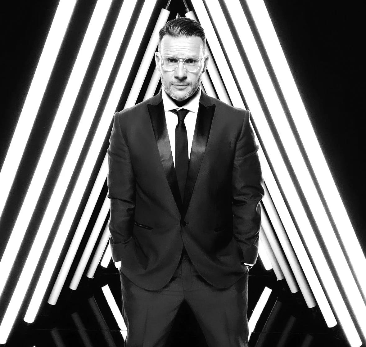 Black and white photograph of Kyle Christy in a tuxedo and glasses standing in front of a chevron of light tubes.