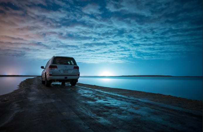 A white car on a causeway road at sunset, the sun setting over still water under streaked clouds.