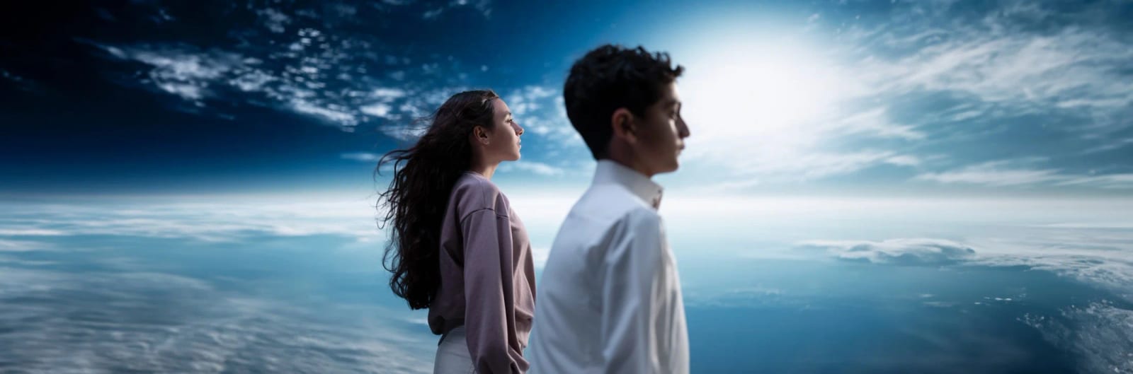 A boy and a girl standing above the clouds looking out over the curve of the Earth, sunlight breaking behind them.
