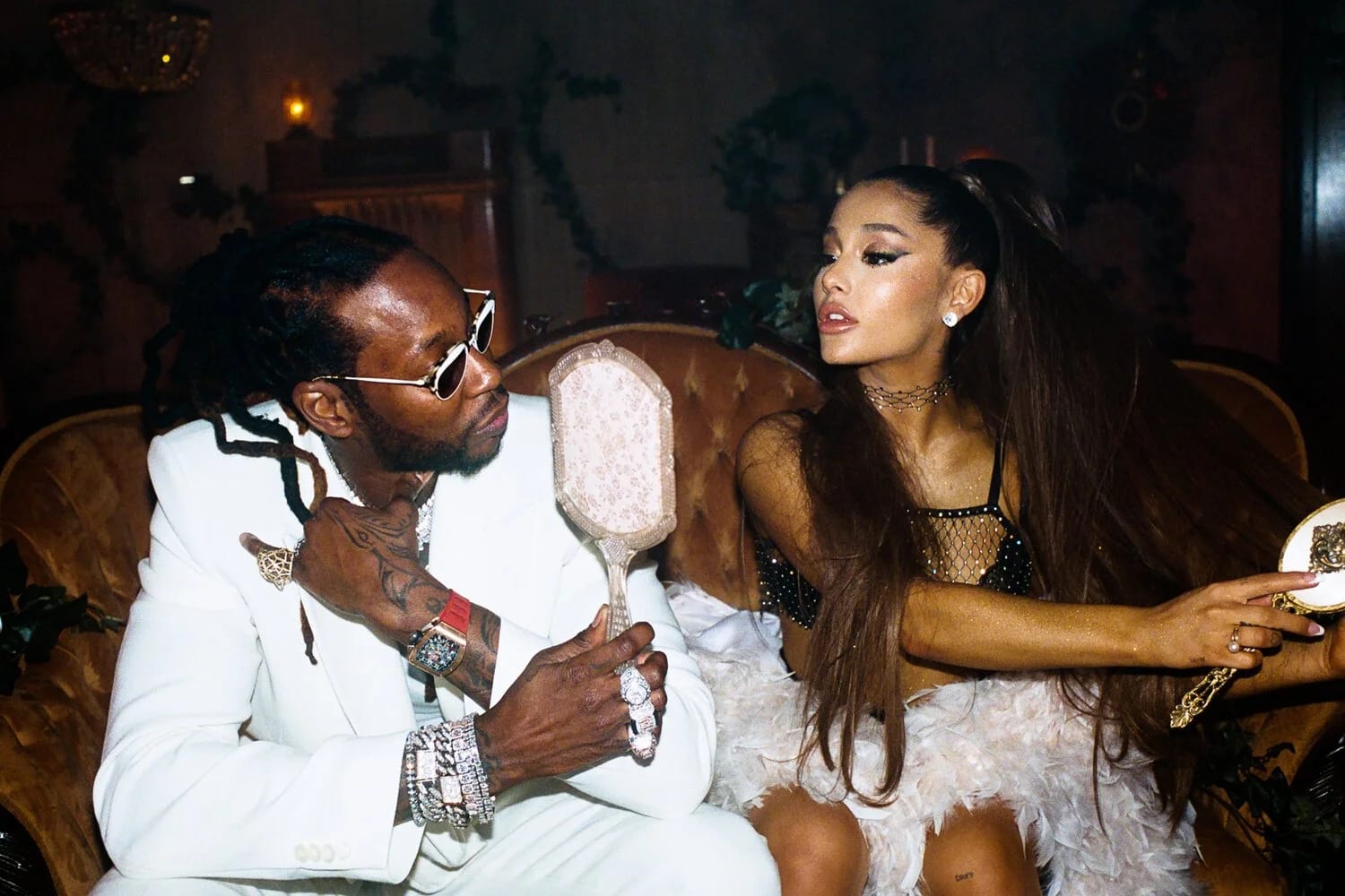 A man in white with sunglasses and a woman with a high ponytail sitting on a velvet sofa, both looking into a hand mirror between them.