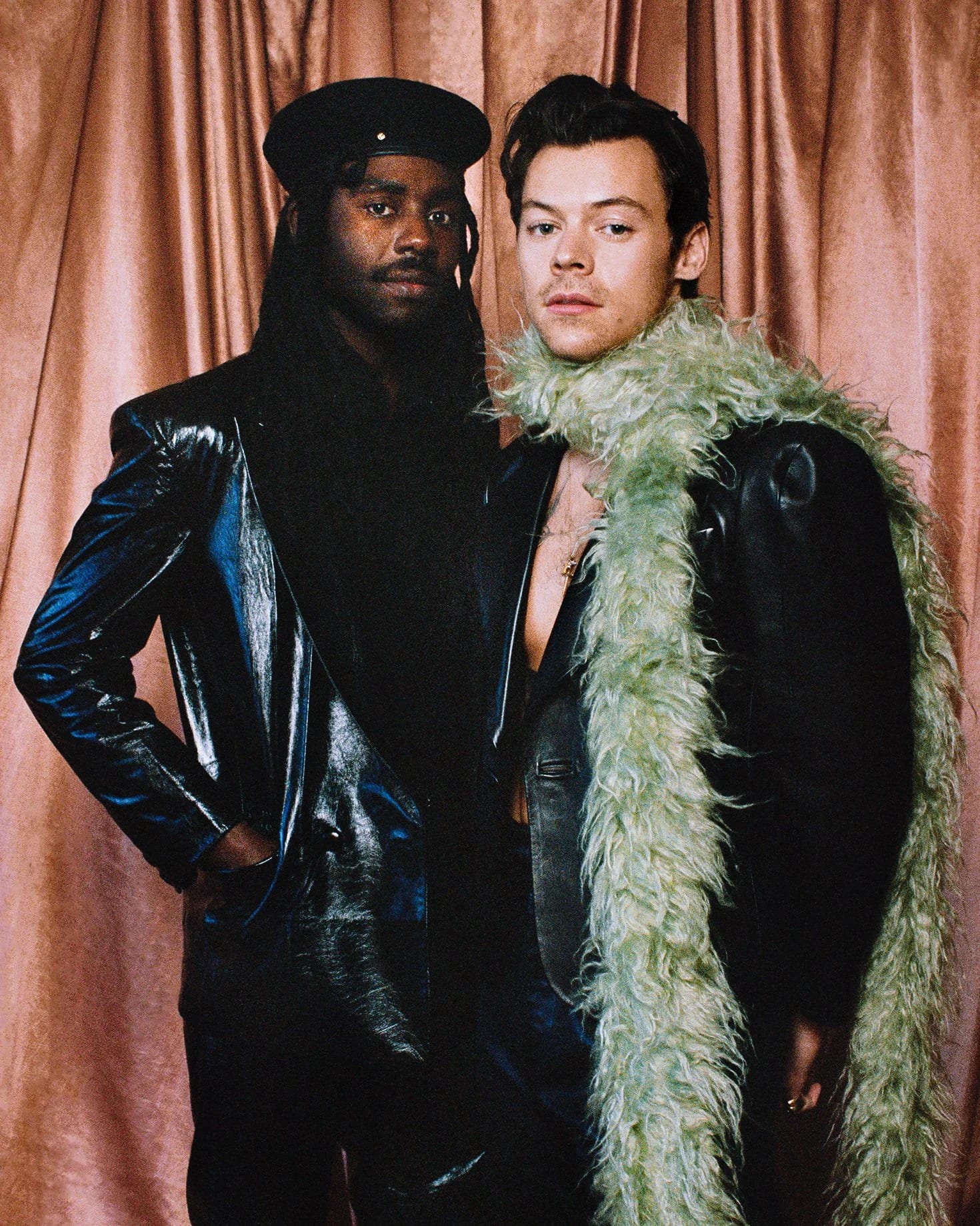 Harry Styles in a leather suit and green feather boa standing beside a man in a black leather suit and cap in front of a pink curtain.