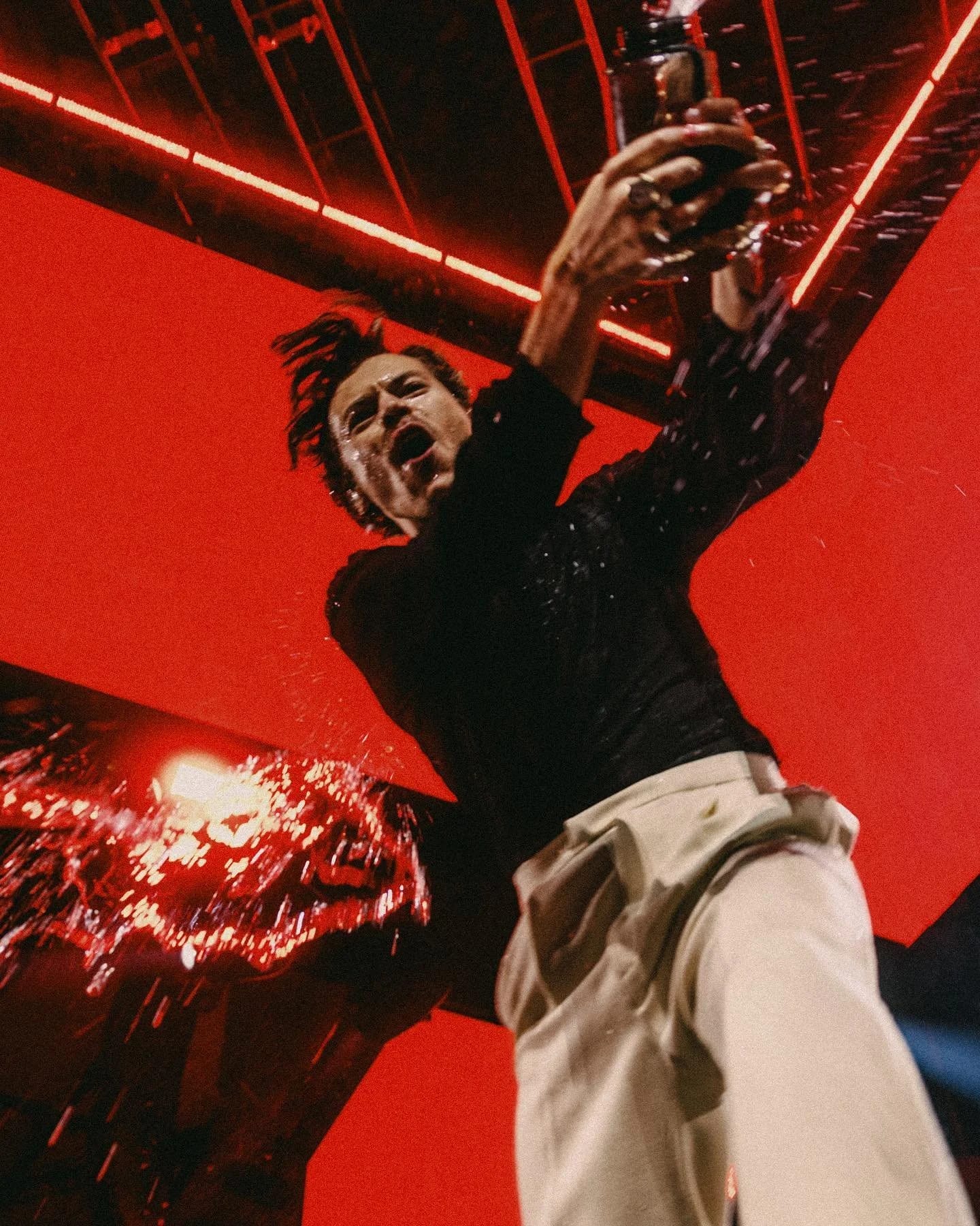 Harry Styles in a black sequinned top and white trousers throwing his hands up under red stage lights.
