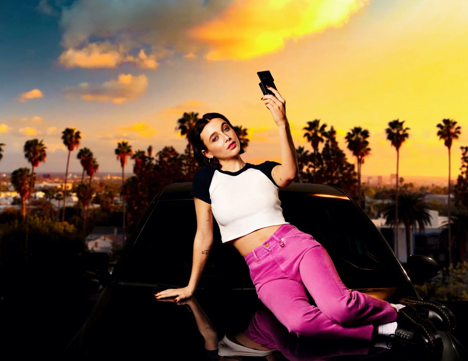 A woman in a white crop top and pink trousers lying on a car bonnet holding up a phone, palm trees and a sunset sky behind.