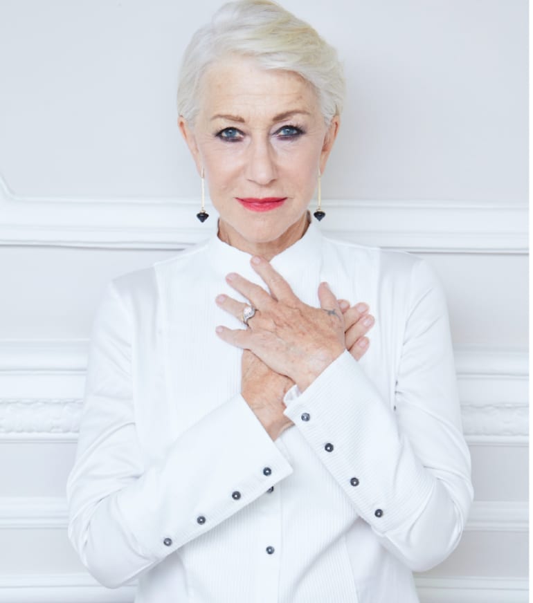An older woman with short platinum hair and long drop earrings, both hands crossed over her chest, in a white shirt with black studs against a white panelled wall.