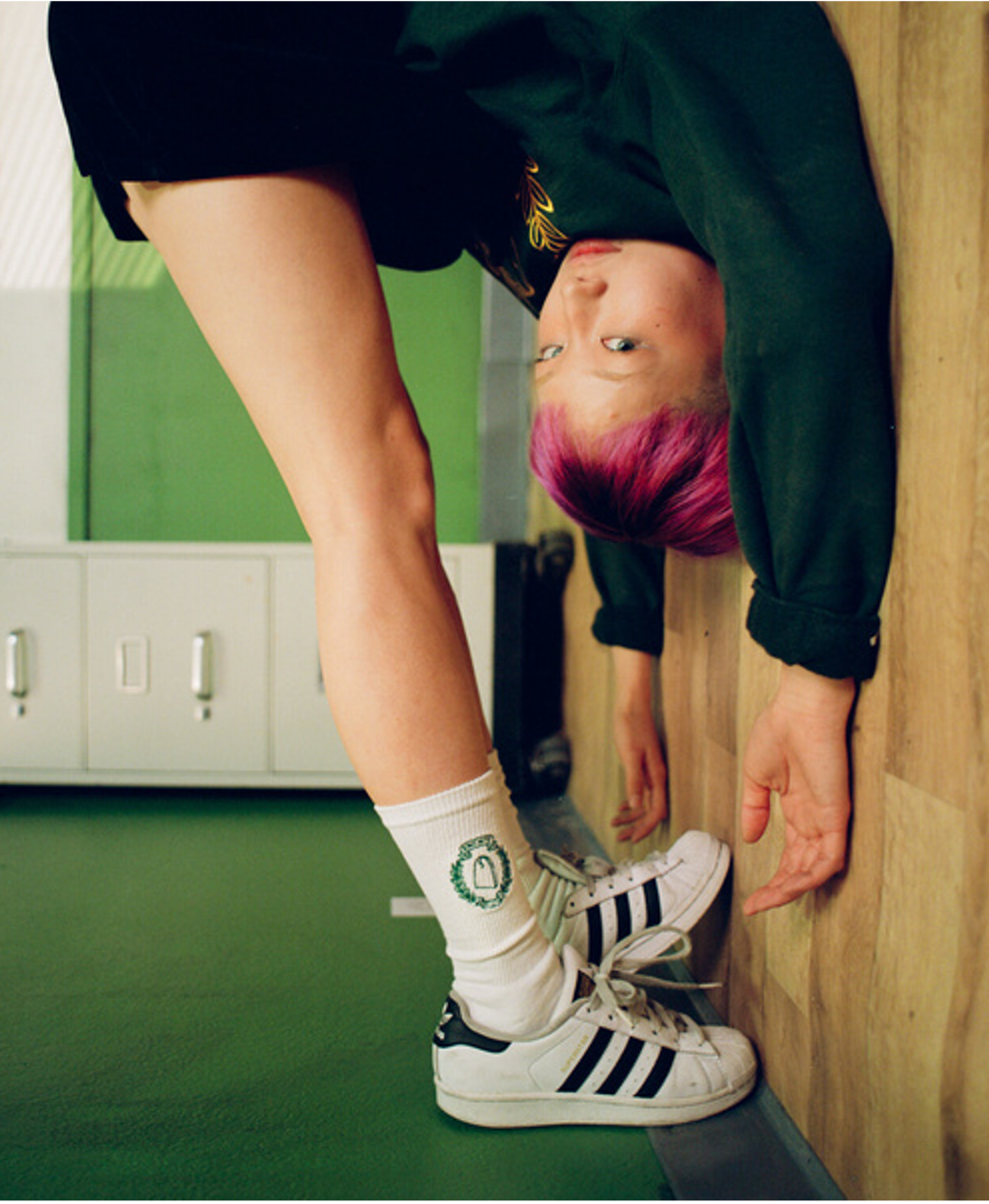 A young woman with pink hair hanging upside down from a bench, her white socks and striped trainers in the foreground.