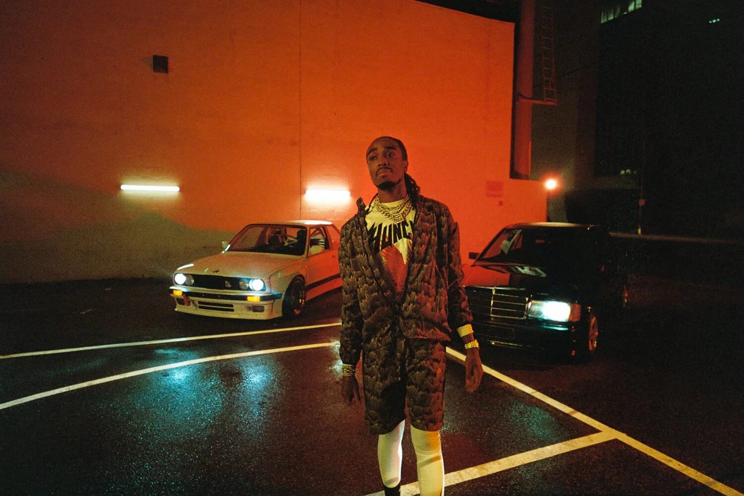 A man in a patterned jacket walking across a car park between two cars, an orange-lit wall behind him.