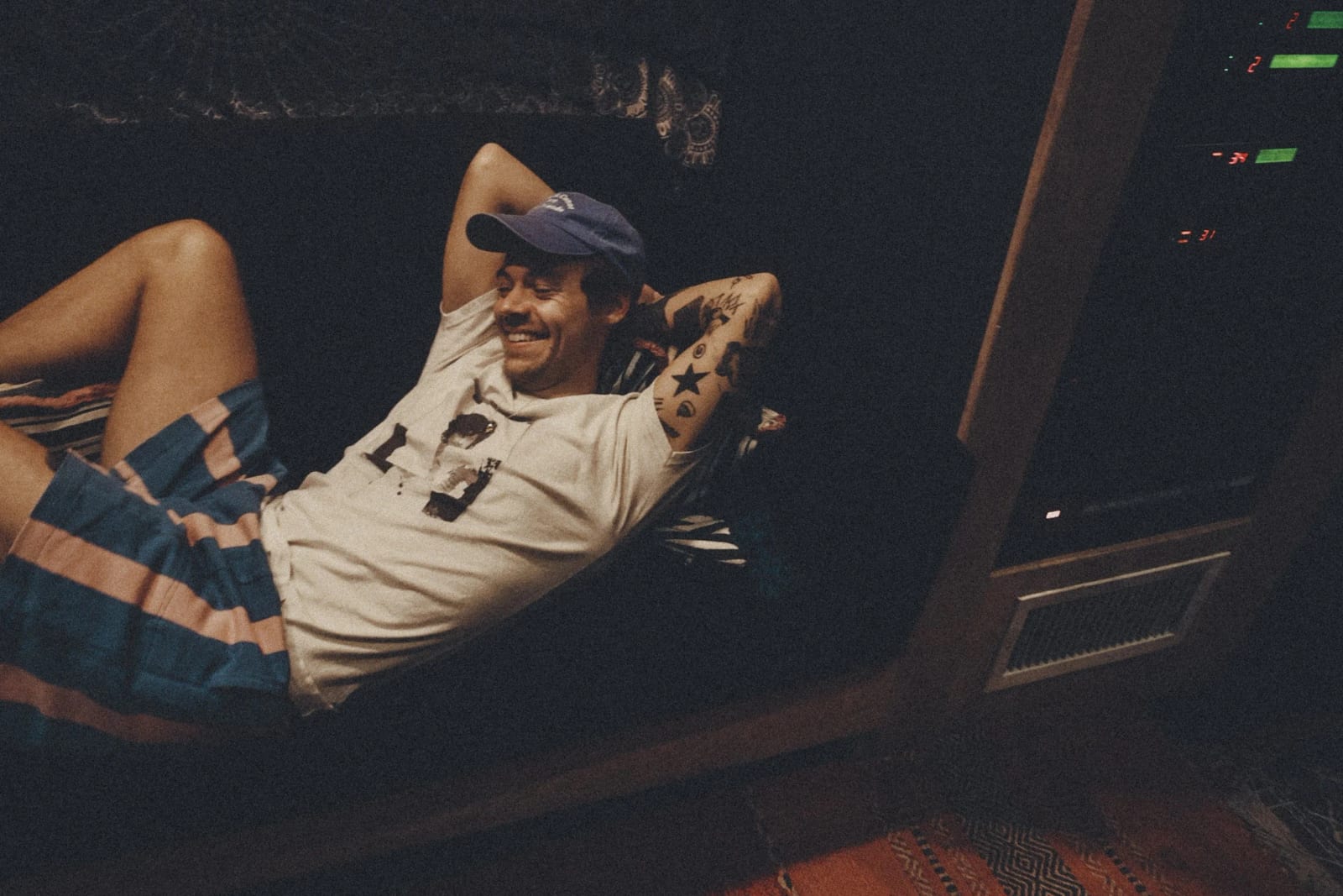 A man in a navy cap and white T-shirt lying back on a striped bus seat with his hands behind his head, grinning.