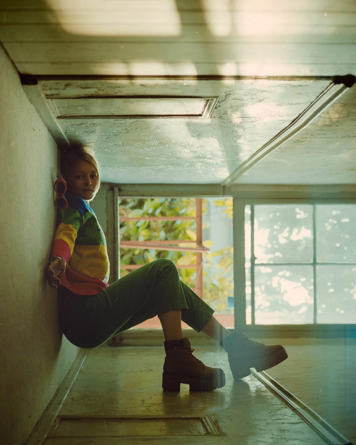 A young woman in a rainbow-striped top and platform boots sitting on the floor of a narrow sunlit corridor, legs stretched out.