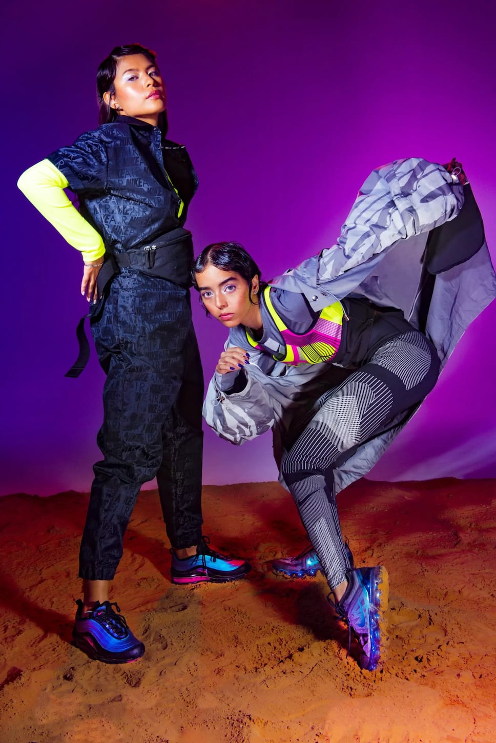 Two women in patterned sportswear and blue trainers posing on sand against purple.