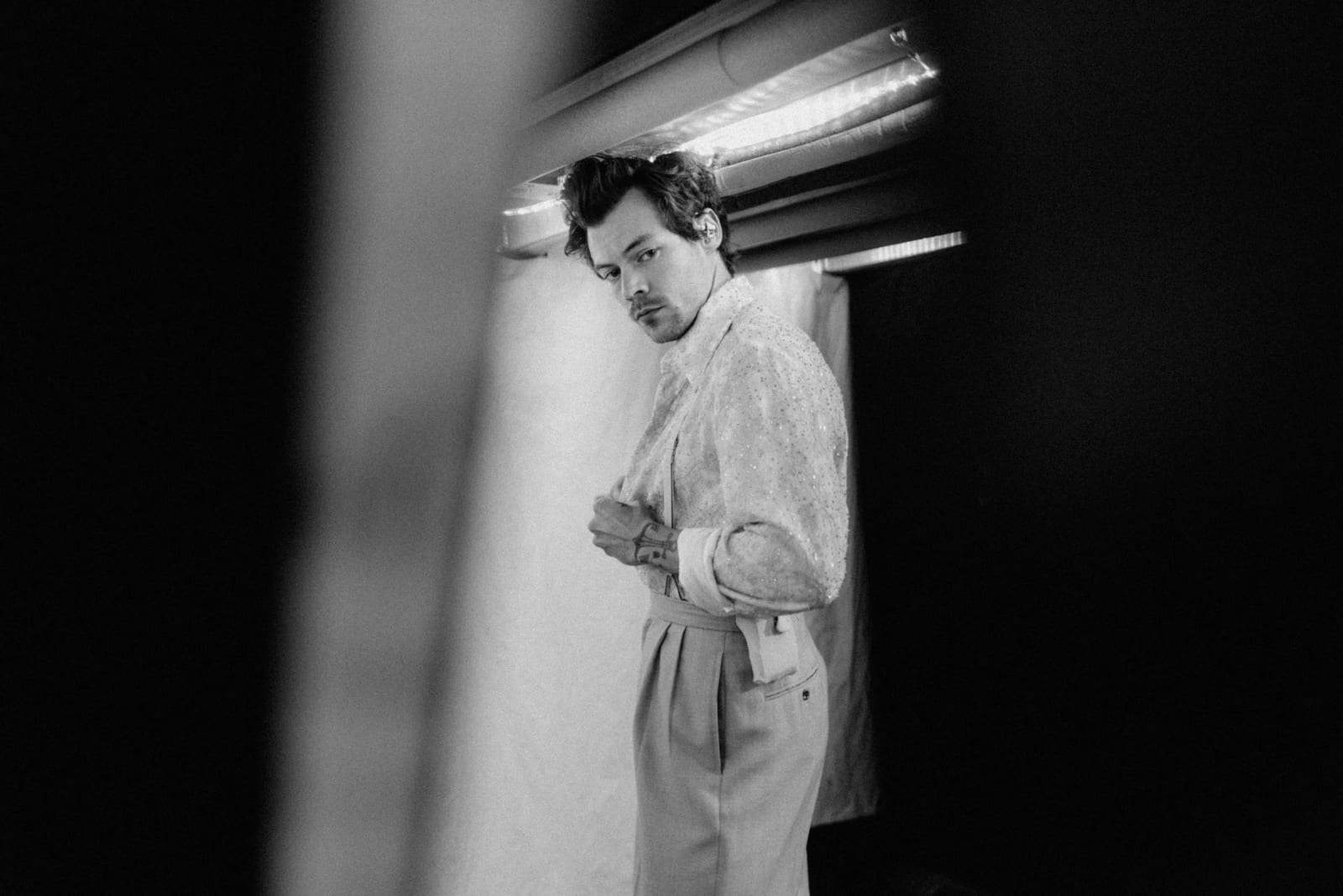 Black and white photograph of a man glancing back over his shoulder in a doorway, buttoning a pale shirt.