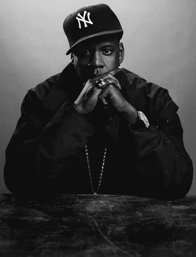Black and white portrait of a man in a New York Yankees cap and dark jacket, hands clasped under his chin.