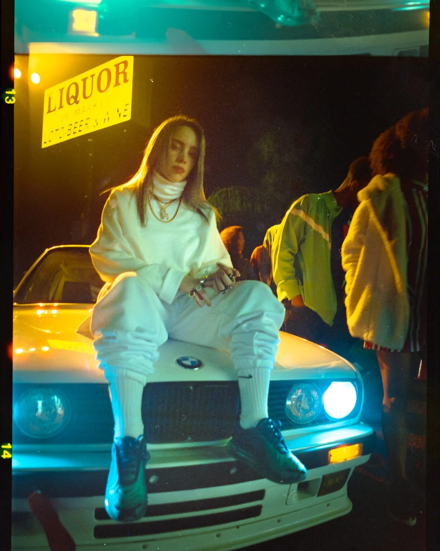 A young woman in an all-white tracksuit sitting on the bonnet of a white BMW at night beneath a yellow LIQUOR sign, people in bright jackets standing to one side.