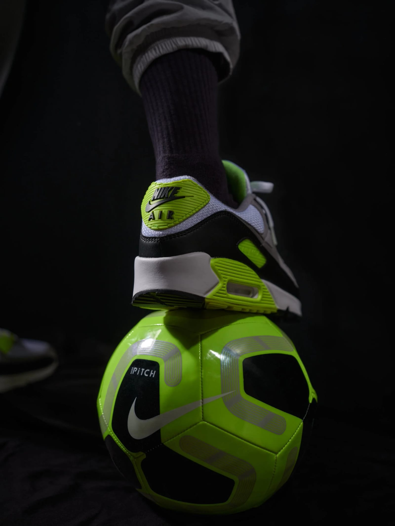 Close-up of a foot in a Nike Air trainer resting on a fluorescent green football in the dark.