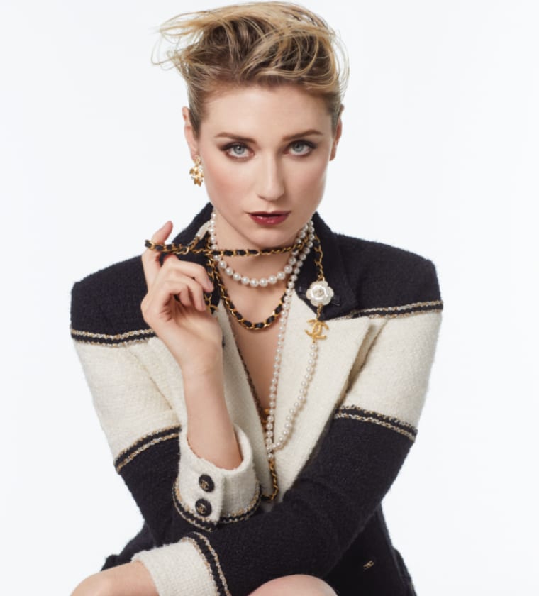A woman with swept-up blonde hair in a black and cream tweed jacket, lifting a beaded necklace to her lips.