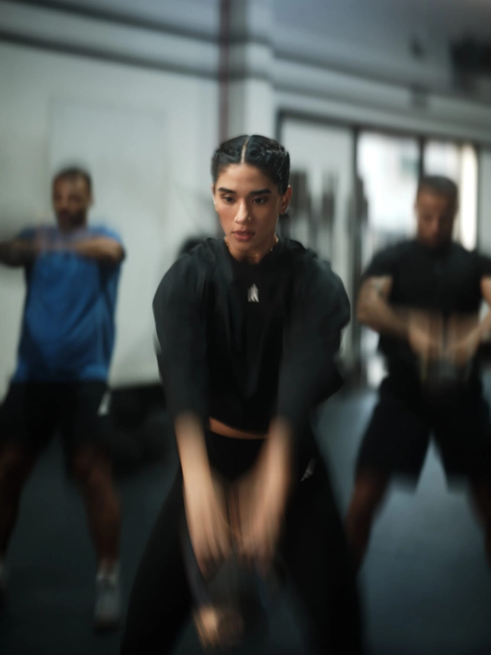 Three people exercising in a gym, the woman in black sportswear at the centre blurred with motion, two men behind her.