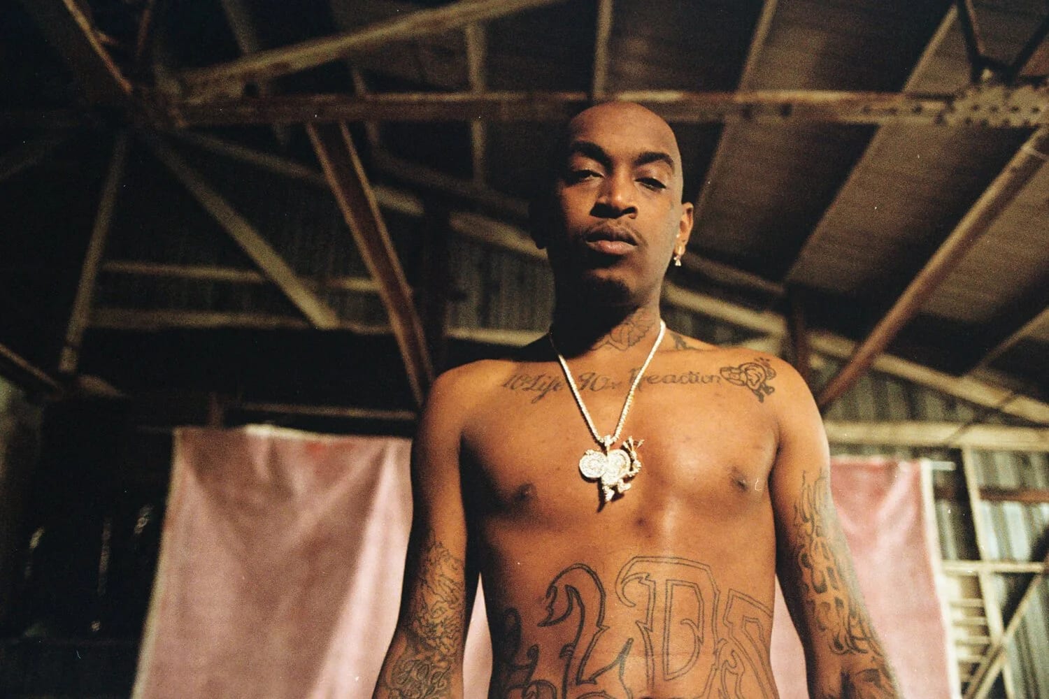 A shirtless man with chest tattoos and a pendant standing in a dilapidated shed, looking down at the camera.