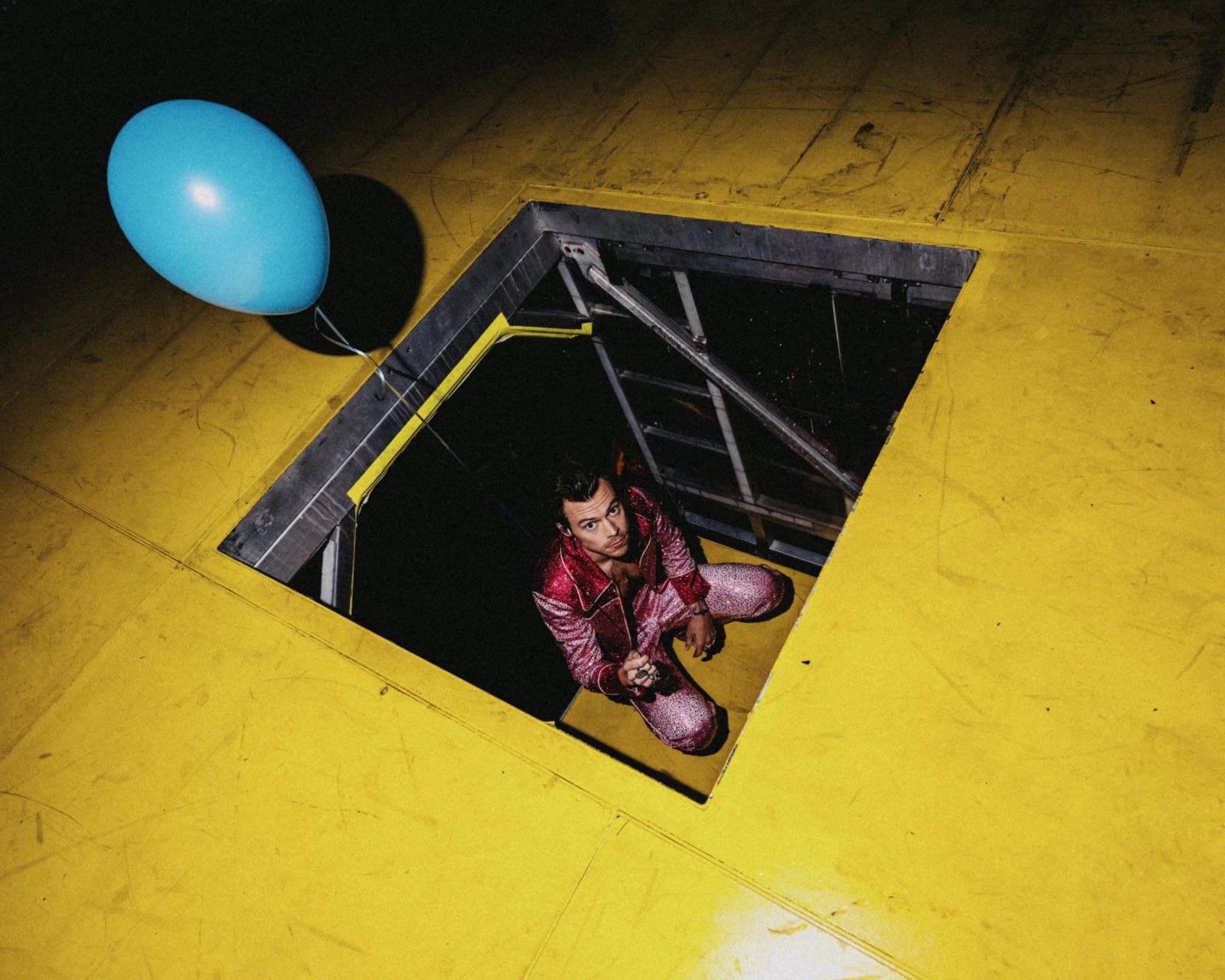 Harry Styles in a pink suit looking up from an open trapdoor in a yellow stage floor, a blue balloon floating beside it.