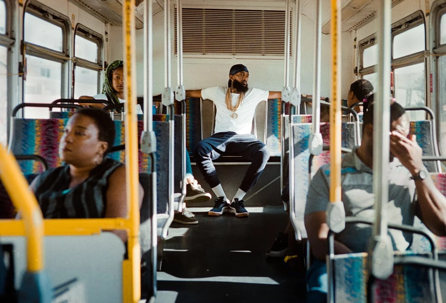 A bearded man in a black durag, white T-shirt and layered gold chains sitting across the back seat of a city bus, arms spread along the seatback, other passengers in the rows ahead.