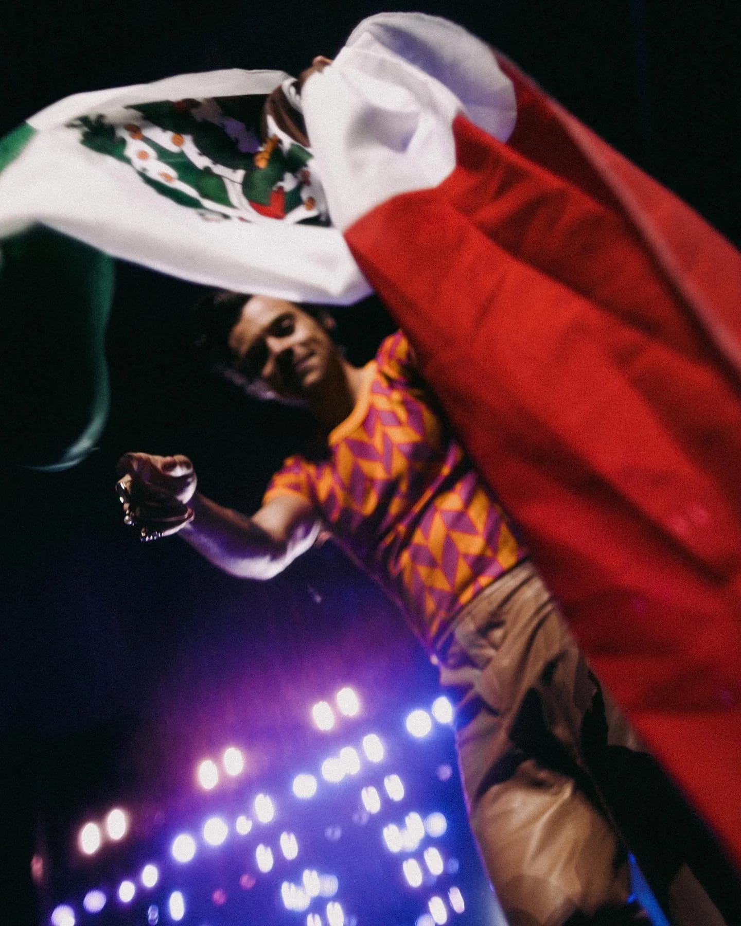 Harry Styles in a patterned top waving a Mexican flag above a sea of stage lights.