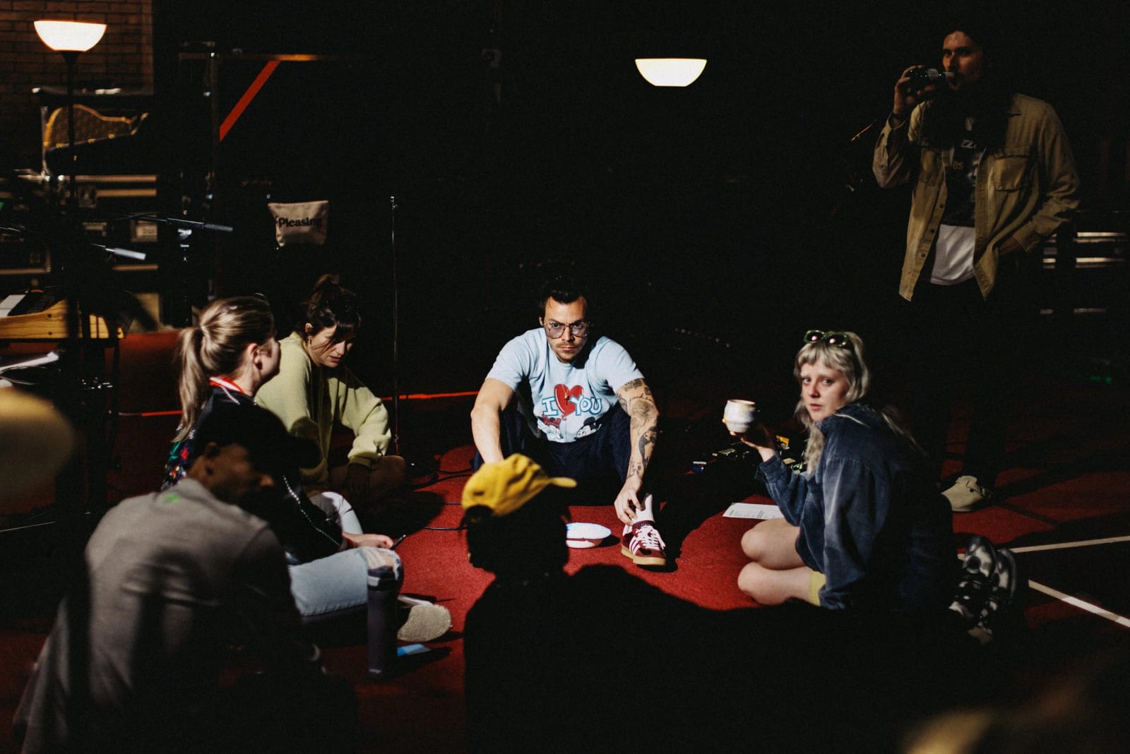 A group of people sitting cross-legged on the floor around a red mat, one man in a white T-shirt at the centre, a photographer standing behind.