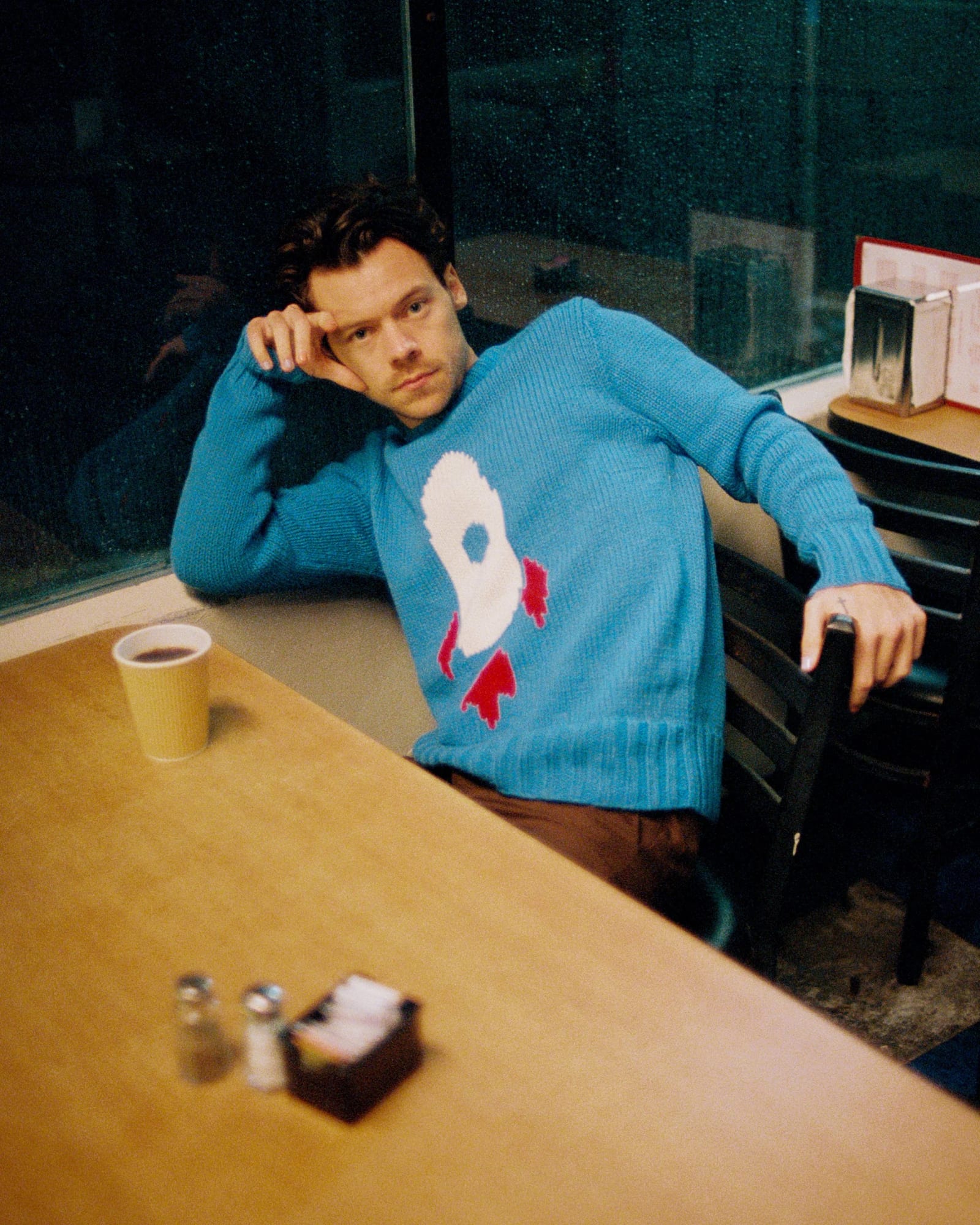 Harry Styles in a blue jumper with a rocket motif leaning back in a diner booth, a paper cup on the table.