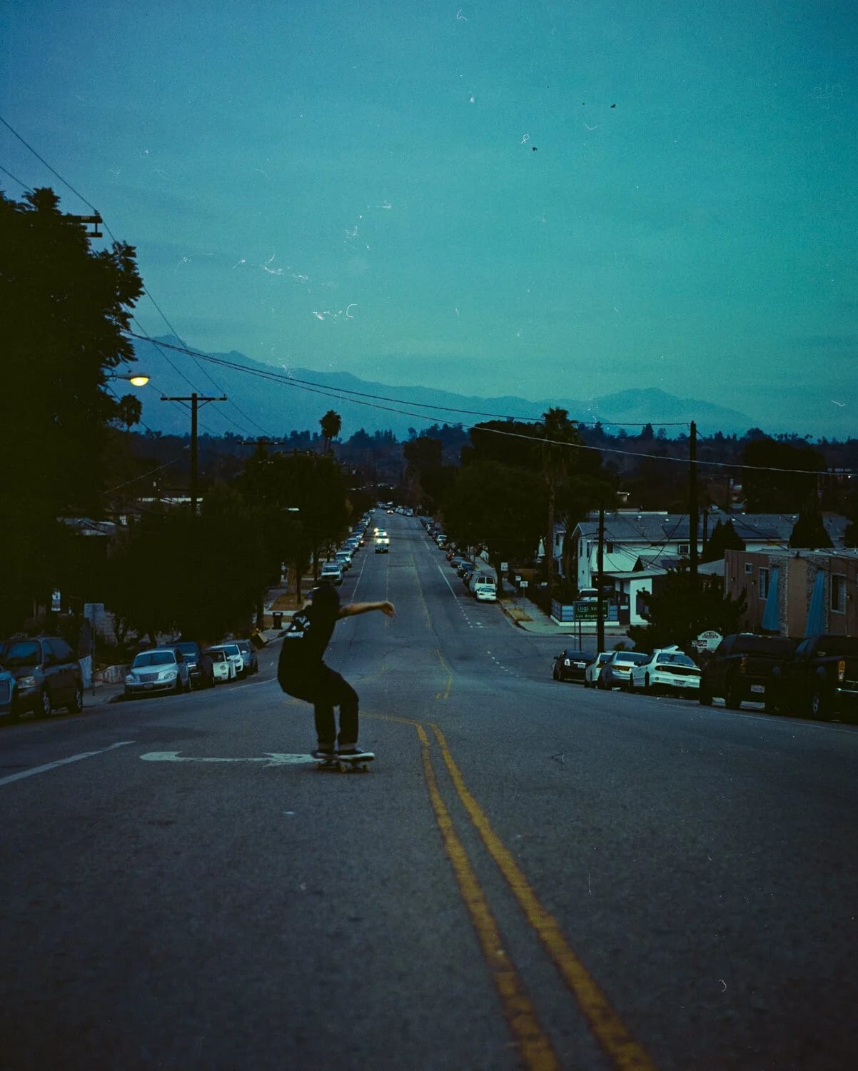 A skateboarder riding down the middle of a residential street at dusk, cars parked either side.